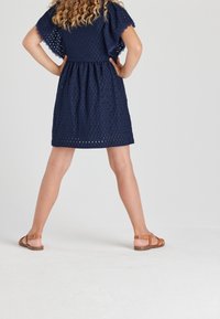 Granatowa sukienka z wycięciami z krótkimi, falbaniastymi rękawami i wcięciem w talii. Połączona z brązowymi sandałami na gołych nogach, na jasnym tle.