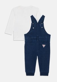Denim overall i mørkeblå med justerbare volangstropper, to baklommer og et trekantet logo-lapp. Overdelen er akkompagnert av en hvit langermet skjorte.