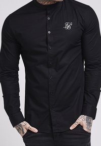 Zwart T-shirt met lange mouwen en een opstaande kraag, voorzien van een knoopsluiting aan de voorkant en een klein wit geborduurd logo op de borst.