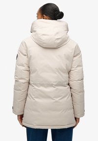 Beżowa kurtka puchowa z wysokim kołnierzem i kapturem, pikowana tekstura, boczne kieszenie oraz naszywka z logo na rękawie.