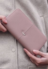 Pink leather wallet with a smooth texture, featuring a silver logo detail. The wallet is held against a gray, textured garment.