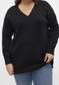 Kobieta w czarnym swetrze z długim rękawem w serek i niebieskich dżinsach, trzymająca brzeg swetra prawą ręką.