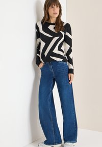 Musta- ja kermahenkinen neulepaita, jossa on siksak-kuvio, yhdistetty leveälahkeisiin sinisiin farkkuihin. Malli seisoo neutraalia taustaa vasten.