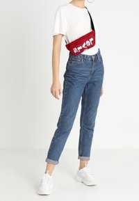 Marsupio rosso in tessuto con testo bianco, indossato a tracolla. Modello in t-shirt bianca a girocollo e jeans blu denim arrotolati, abbinati a sneakers bianche.