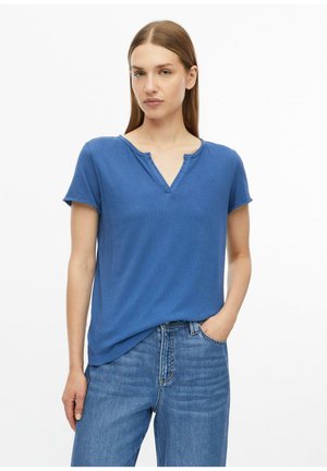 Femme aux cheveux lisses châtain clair porte un haut bleu à manches courtes et col en V ainsi qu'un jean bleu, debout avec une main dans la poche sur fond blanc.