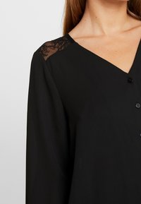 Czarna bluzka z dekoltem w kształcie V, z zapięciem na guziki i koronkowymi detalami na rękawach, wykonana z gładkiego materiału.
