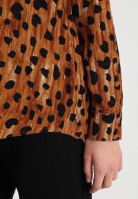 Blusa marrone con un motivo a leopardo nero, dotata di polsini con bottoni e tessuto testurizzato, abbinata a un tessuto nero uniforme sotto.