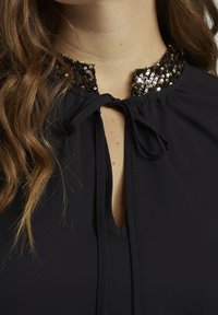 Svart blus med en halsringning prydd med paljetter och knytning, mjukt tyg och nyckelhålsdesign. Slät textur och elegant detalj.