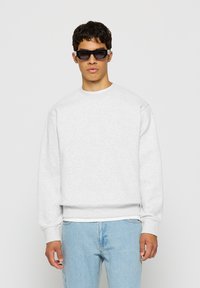 Young man wearing black rectangular sunglasses, a light gray sweatshirt, and light blue jeans, standing against a plain white background.