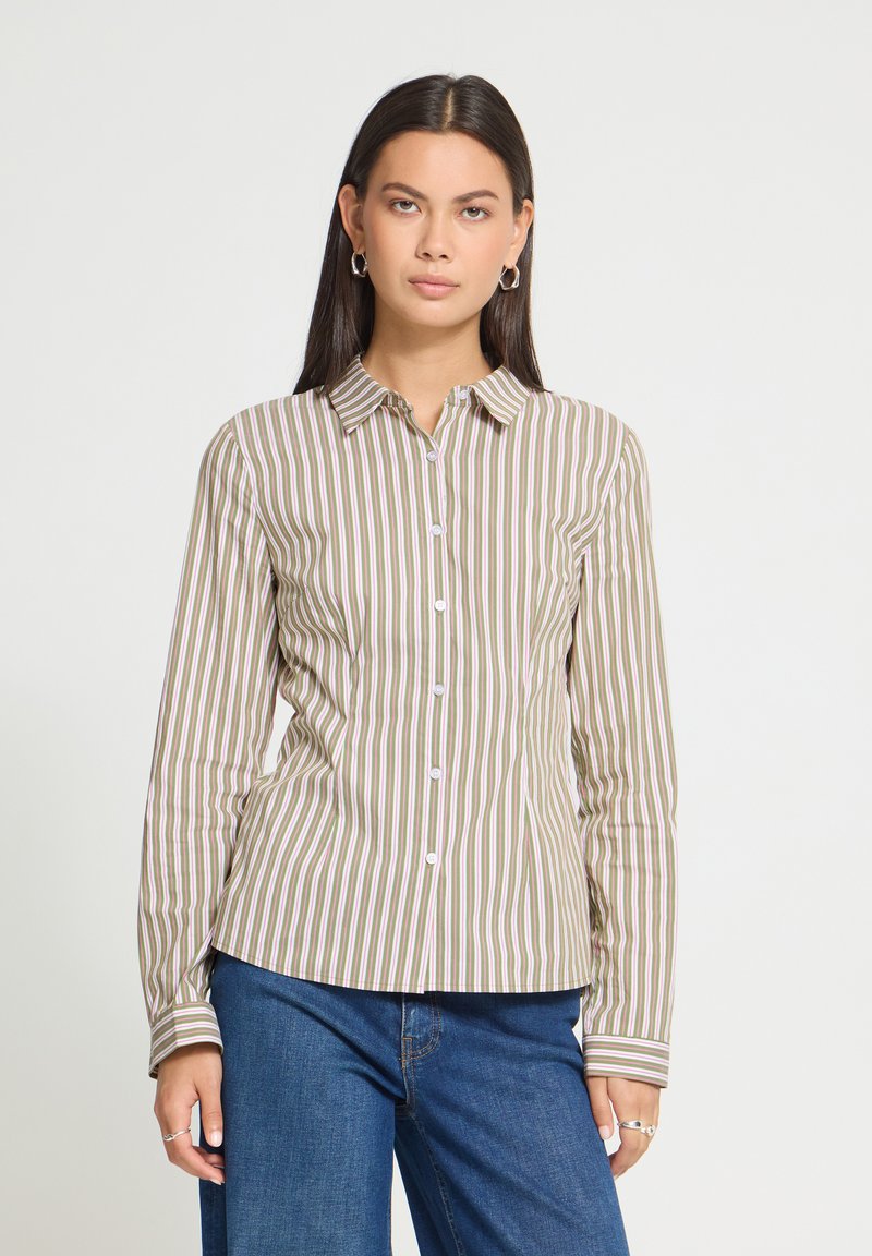 Femme portant une chemise beige et blanche à rayures verticales boutonnée, manches longues, et un jean bleu, debout devant un fond clair uni.