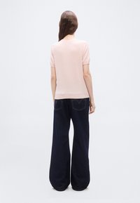 Μια γυναίκα στέκεται με την πλάτη στραμμένη προς τα εμπρός, φορώντας ένα ανοιχτό ροζ κοντομάνικο τοπ και σκούρα φαρδιά τζιν παντελόνια, μπροστά σε ένα απλό φόντο.