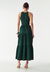 Temno zelena maxi obleka s tiered dizajnom, ki vsebuje prilega bodice, naramnice in nabrano pas. Gladek, teksturiran material.