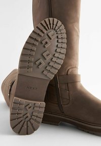 Bottes marron mi-mollet avec semelles en caoutchouc robustes, fermeture éclair latérale et sangle décorative avec détail de boucle, présentées sur un fond uni.