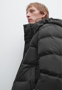 Schwarze Pufferjacke mit hohem Kragen, ausgestattet mit horizontal gequilteten Abschnitten und großen schwarzen Knöpfen. Glatte, glänzende Textur, minimalistisches Design.