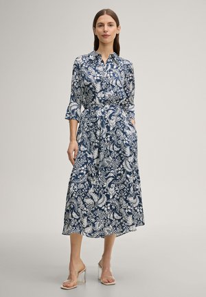 Vrouw staand in een blauw-witte midi-jurk met bloemen, 3/4 mouwen en transparante sandaal met blokhak tegen een effen achtergrond.