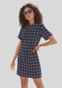 Navyblaues Kurzarmkleid mit kariertem Muster, aus strukturiertem Stoff, mit taillierter Passform und Taschen. Kombiniert mit Sonnenbrille.