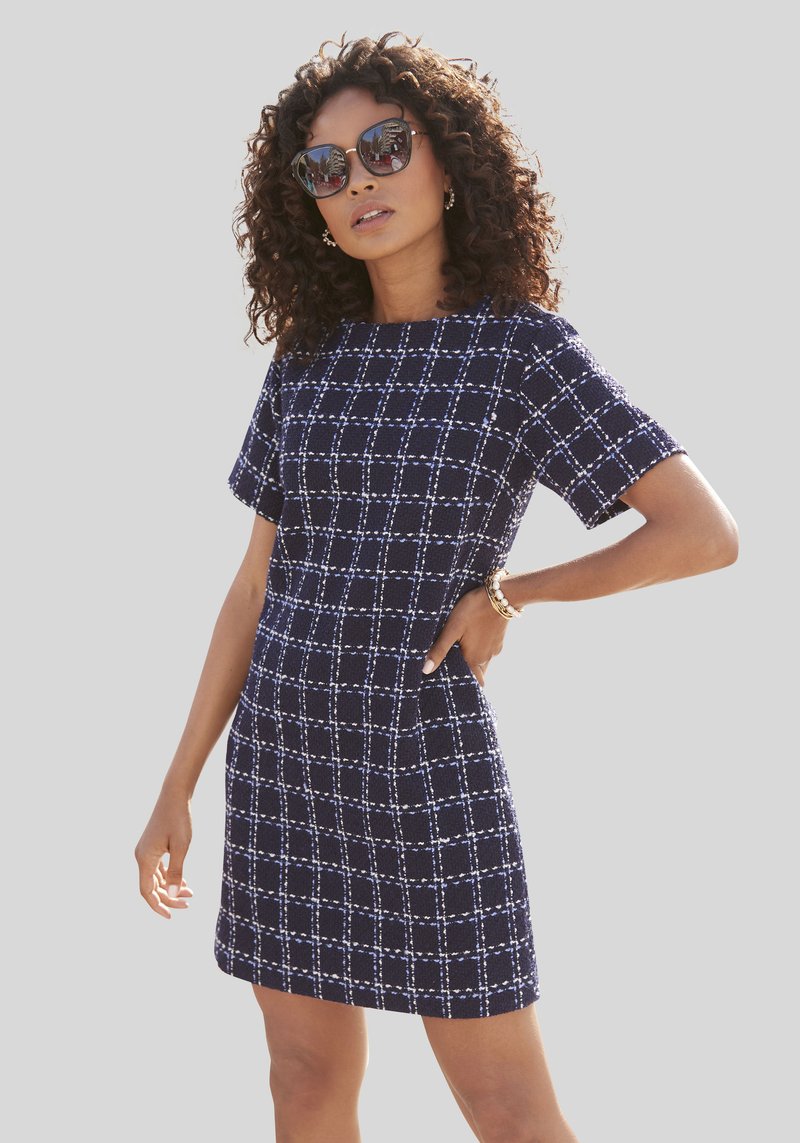 Navyblaues Kurzarmkleid mit kariertem Muster, aus strukturiertem Stoff, mit taillierter Passform und Taschen. Kombiniert mit Sonnenbrille.