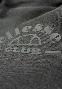 Textile gris avec une texture douce, orné de motifs imprimés dans une teinte plus claire. Le texte indique "Essentials Club" avec un logo ressemblant à une forme de dôme.