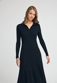 Svart ribbad klänning med V-ringning och kragdesign, långa ärmar, flytande siluett och en texturerad tygfinish.