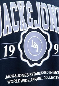 Marineblauwe t-shirt met grote witte en lichtblauwe tekst; embleem met "J&J" logo, jaartal "1990," en merkinformatie eronder gedrukt.