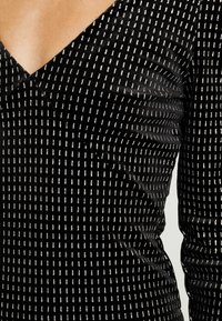 Svart tygblus med V-ringning, långa ärmar och ett strukturerat mönster av silverfärgade horisontella linjer. Slätt material med lätt glans.