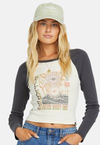 Korte mouwen cropped shirt in crème en donkergrijs met een grafische print van ogen en golven. Gecombineerd met een groene Billabong pet.