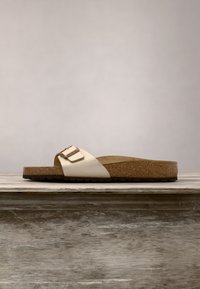 Sandale à enfiler en cuir beige avec semelle en liège marron et boucle en or rose sur une surface en bois sur fond uni.