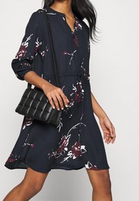 Marineblauwe bloemenjurk met lange mouwen, knoopsluiting en strik taille, gecombineerd met een zwart gewatteerde schoudertas. Glad stoffen textuur.