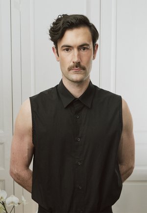 Man met donker golvend haar en snor, gekleed in een mouwloos zwart overhemd met knopen, staand tegen een witte muur met handen achter de rug.