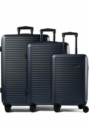 Tre valigie rigide blu scuro con scanalature orizzontali, maniglie telescopiche e quattro ruote girevoli nere, disposte per dimensione.