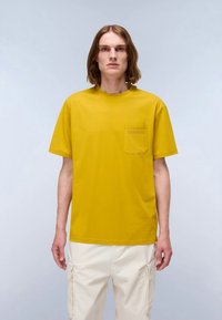 Mosterdgele katoenen T-shirt met een ronde halslijn en korte mouwen. Heeft een borstzak aan de linkerkant met een gestikt detail.