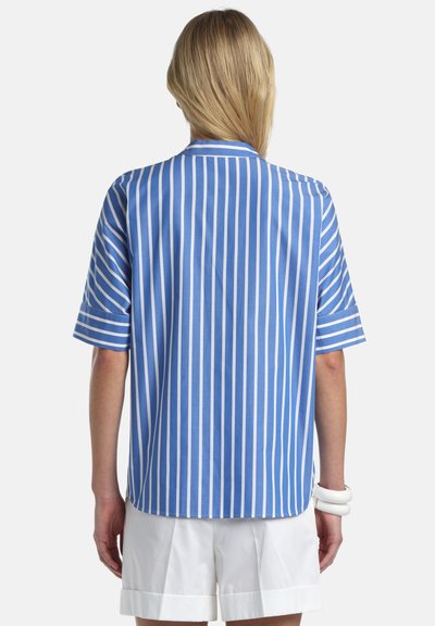 Personne blonde vue de dos portant une chemise à manches courtes rayée verticalement bleu et blanc, un short blanc à revers et des bracelets blancs.