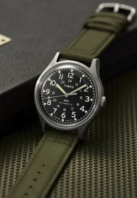 Analógové automatické hodinky Timex MK-1 s čiernym ciferníkom, bielymi číslicami a zeleným látkovým remienkom na textúrovanom zelenom povrchu a tmavej krabičke.