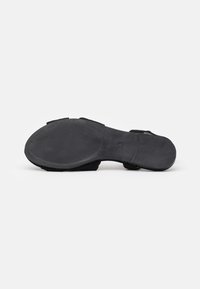 Semelle de sandale noire vue de dessous, montrant un motif de grip texturé à l'avant et une zone de talon lisse sur un fond blanc uni.