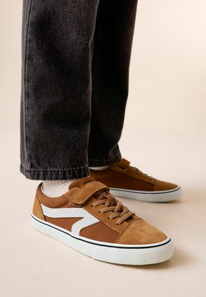 Brune sneakers med hvide accenter, snørebånd og en velcrolukning, båret med sorte jeans og hvide sokker på en lys baggrund.