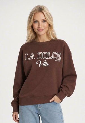 Blonde vrouw die een bruine trui draagt met de tekst "LA DOLCE Vita" en lichtblauwe jeans, staand met één hand in haar zak, glimlachend.