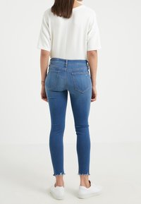 Jeans skinny de mezclilla azul con dobladillo deshilachado, cintura alta y bolsillos traseros, combinados con una blusa blanca de manga corta y zapatillas blancas.