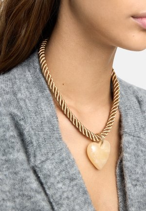 Złoty naszyjnik z plecionym sznurem i dużym beżowym sercem jako zawieszką, noszony na szarym dzianinowym kardiganie.