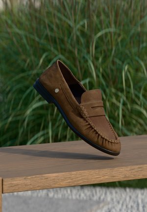 Mocassino da uomo in camoscio marrone con suola nera appoggiato su una panchina di legno, con erba alta verde sfocata sullo sfondo.