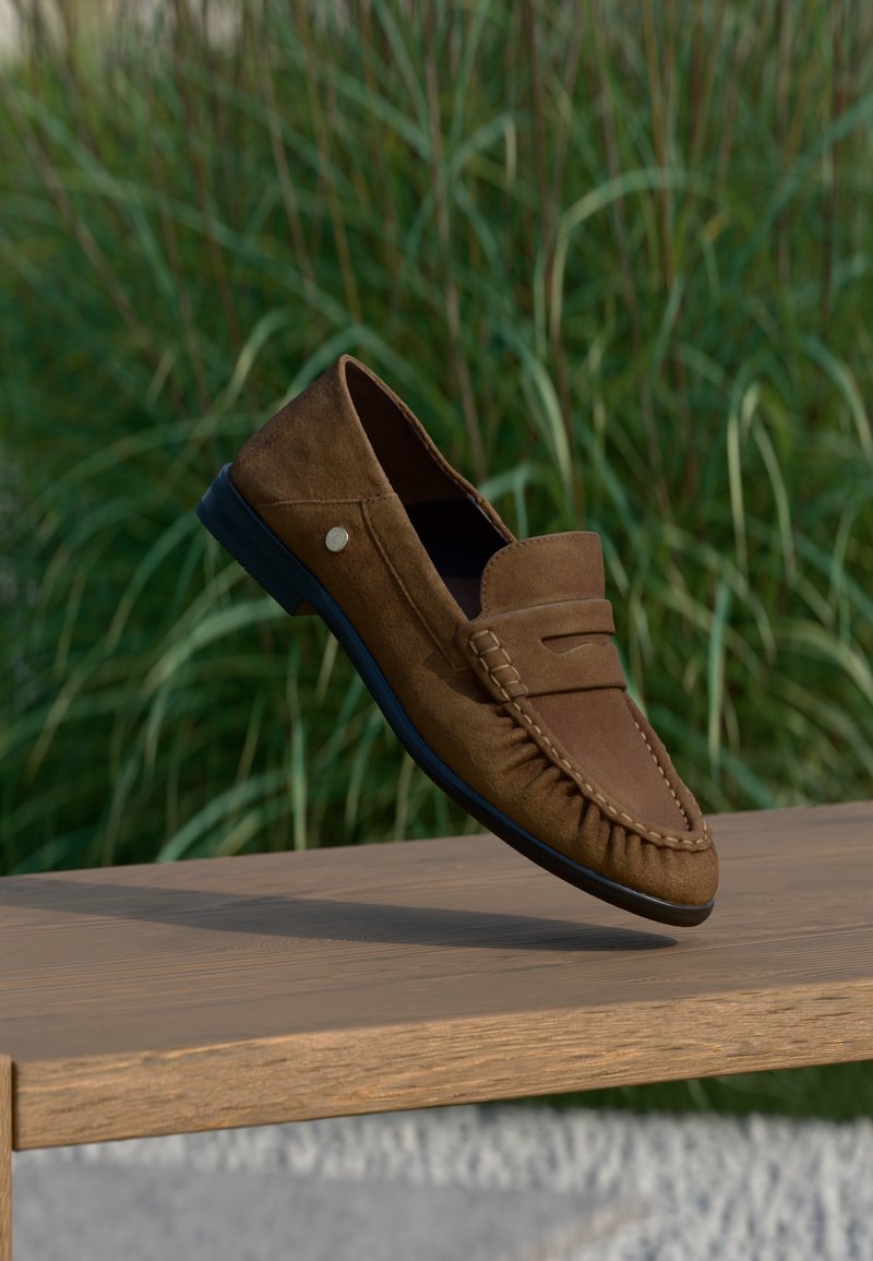 Καφέ δερμάτινη ανδρική μοκασίνη με μαύρη σόλα ισορροπημένη σε ξύλινο παγκάκι, πράσινο ψηλό γρασίδι θολό στο φόντο.