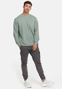 Lys grøn langærmet sweatshirt med en brystlomme og logo-prydet halsudskæring, parret med grå cargobukser og hvide sneakers.
