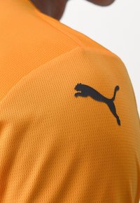 Camisa deportiva naranja con una tela texturizada y transpirable. Presenta un logotipo negro de Puma en el hombro.
