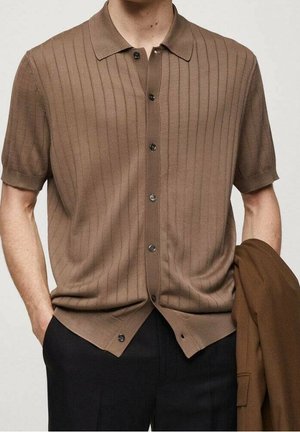 Korte mouw bruin button-up gebreid shirt, ongeknoopt, met zwarte broek, één hand in de zak, de andere houdt een bruine jas vast.