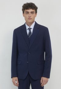 Marineblaues Nadelstreifenanzug mit zwei Knöpfen, Reverskragen und aufgesetzten Taschen. Kombiniert mit einem hellblauen gestreiften Hemd und einer marineblauen Krawatte mit goldenen Akzenten.