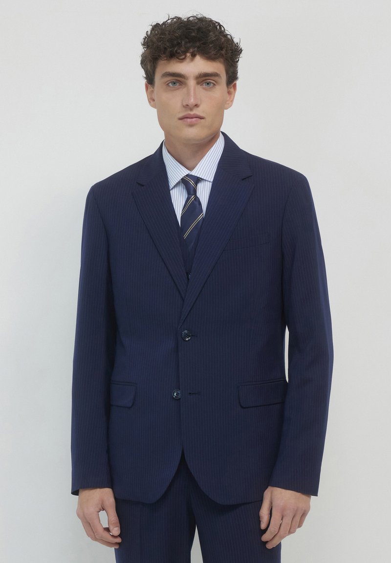 Marineblaues Nadelstreifenanzug mit zwei Knöpfen, Reverskragen und aufgesetzten Taschen. Kombiniert mit einem hellblauen gestreiften Hemd und einer marineblauen Krawatte mit goldenen Akzenten.