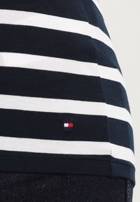 Bawełniany T-shirt w granatowo-białe paski o dopasowanym kroju. Na dole po lewej stronie znajduje się małe logo w kolorach czerwonym, białym i niebieskim.