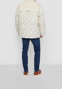 Jasbeżowa kurtka puchowa z wzorzastą dolną częścią, kremowymi ramionami i elastycznymi mankietami, w zestawie z niebieskimi dżinsami i brązowymi butami.