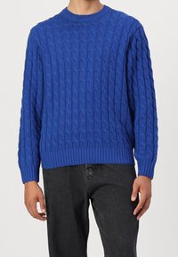 Blauer gestrickter Pullover mit strukturiertem Zopfmuster, rundem Halsauschnitt sowie gerippten Bündchen an Ärmeln und Saum. Getragen mit dunkelgrauer Hose.