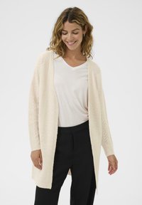 Lång, öppen framsideskofta i mjuk beige stickad med texturerat zigzagmönster. Bärs över en vit V-ringad skjorta och svarta byxor.