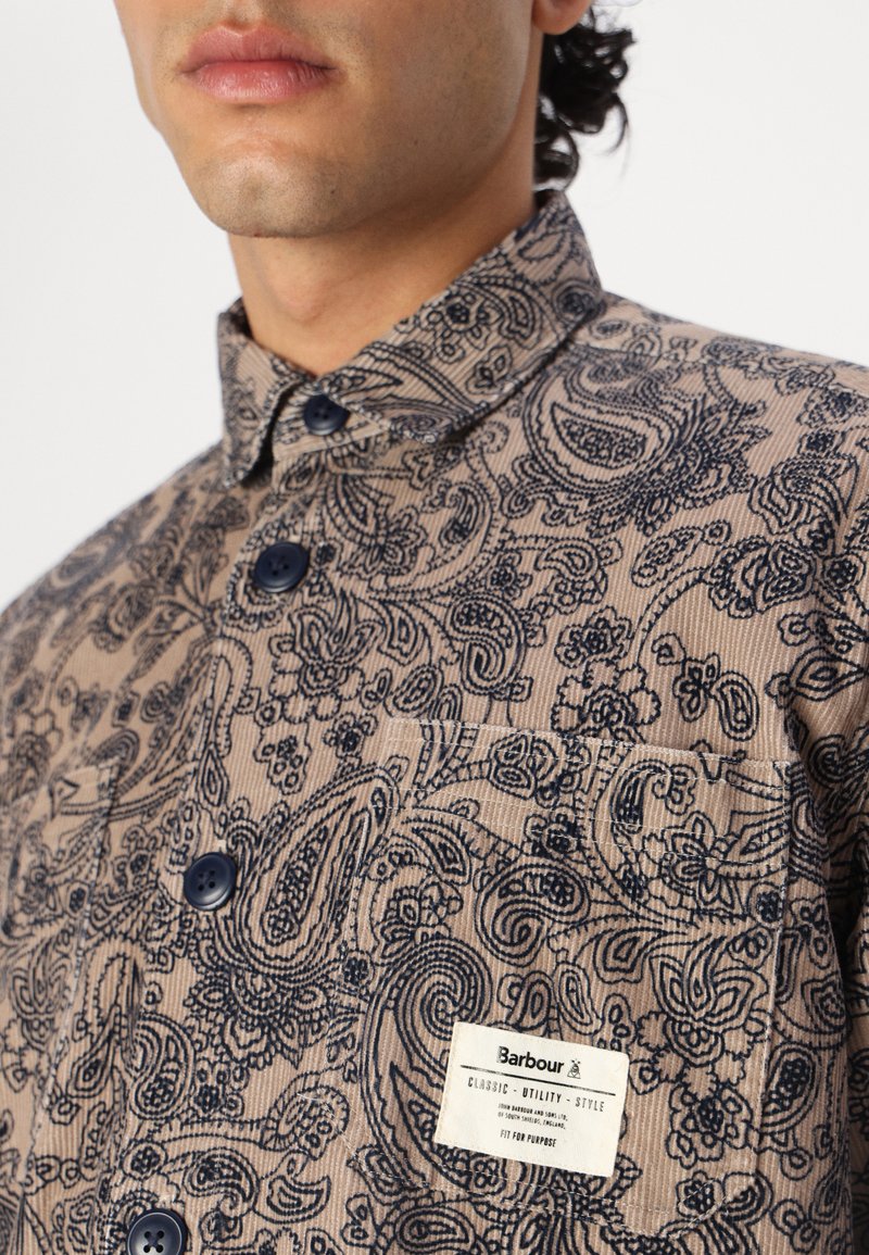 Chemise en velours côtelé beige avec un motif paisley bleu marine, grands boutons, col droit et une poche poitrine avec une étiquette de marque.
